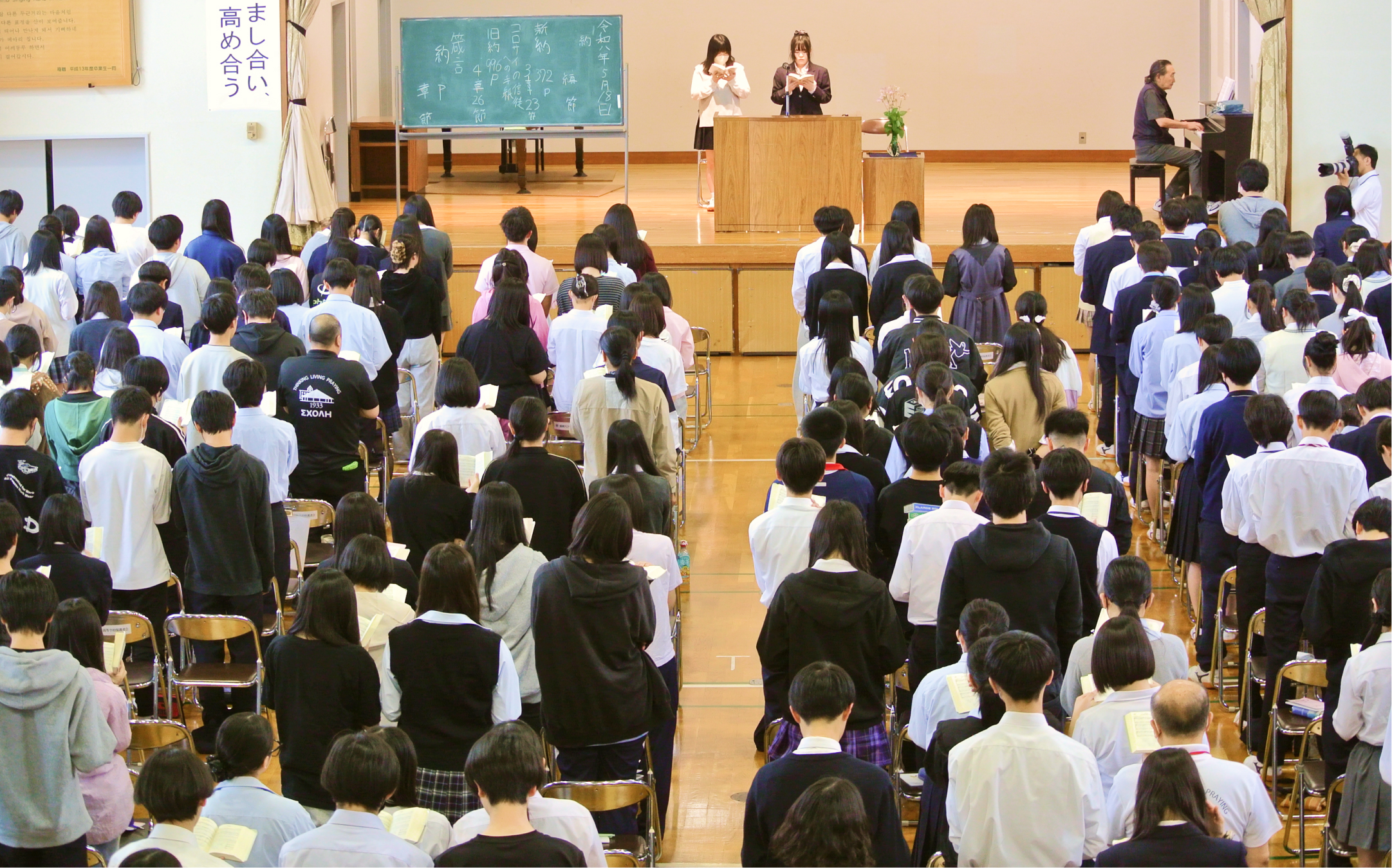 体育館で立って聖書を読む学生たちの写真（大）